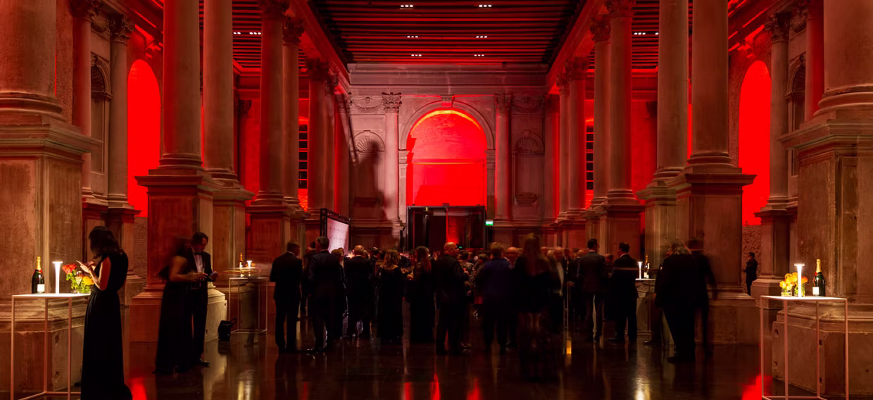 A red architectural interior filled with people gathered in conversation, warm light and spatial depth.