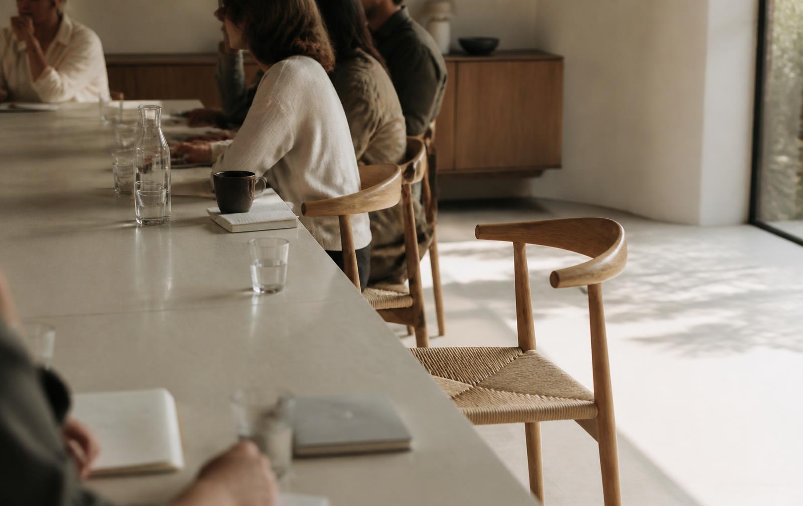 An empty chair beside occupied seats at a long meeting table in daylight.