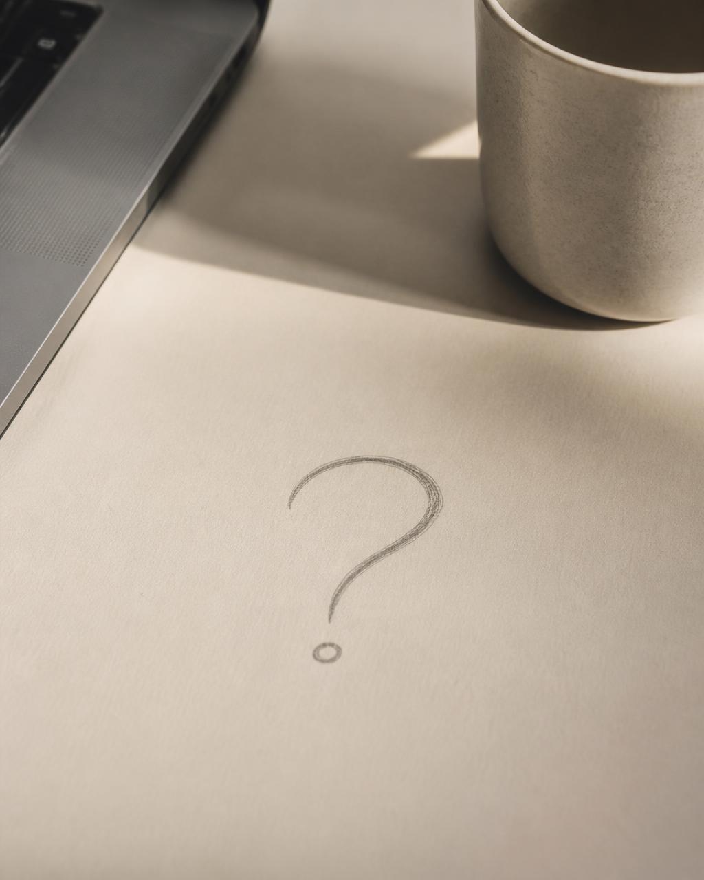 A question mark pencilled on paper beside a laptop and a cup, soft daylight.